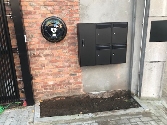 Zwarte modulaire wandbrievenbus met vier postvakken en codepaneel, gemonteerd op een bakstenen muur.