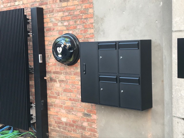 Zwarte modulaire wandbrievenbus met vier postvakken en codepaneel, gemonteerd op een bakstenen muur.