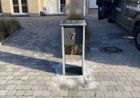 Screenshot 2025-12-15 162213 Zwarte modulaire wandbrievenbus met vier postvakken en codepaneel, gemonteerd op een bakstenen muur.