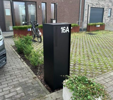 Screenshot 2025-12-15 162914 Zwarte modulaire wandbrievenbus met vier postvakken en codepaneel, gemonteerd op een bakstenen muur.